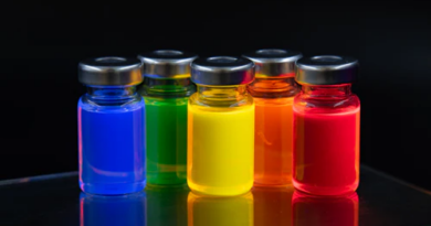 Five glass vials containing fluorescent liquids in blue, green, yellow, orange, and red, glowing under UV light on a reflective surface