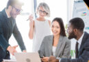 A diverse group of business professionals discussing ideas around a laptop, smiling and collaborating in a modern office setting.