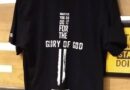 Black T-shirt with a religious-themed design on the back, featuring a vertical white cross integrated with text that reads, "Whatever you do, do it for the glory of God," along with a Bible verse reference (1 Corinthians 10:31). The T-shirt is displayed on a wooden hanger against a wooden background.