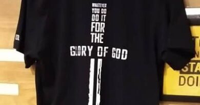 Black T-shirt with a religious-themed design on the back, featuring a vertical white cross integrated with text that reads, "Whatever you do, do it for the glory of God," along with a Bible verse reference (1 Corinthians 10:31). The T-shirt is displayed on a wooden hanger against a wooden background.