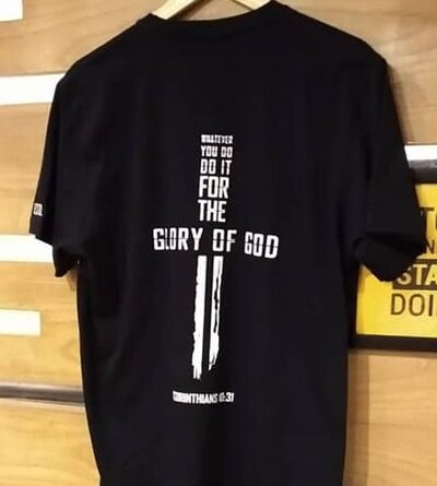 Black T-shirt with a religious-themed design on the back, featuring a vertical white cross integrated with text that reads, "Whatever you do, do it for the glory of God," along with a Bible verse reference (1 Corinthians 10:31). The T-shirt is displayed on a wooden hanger against a wooden background.